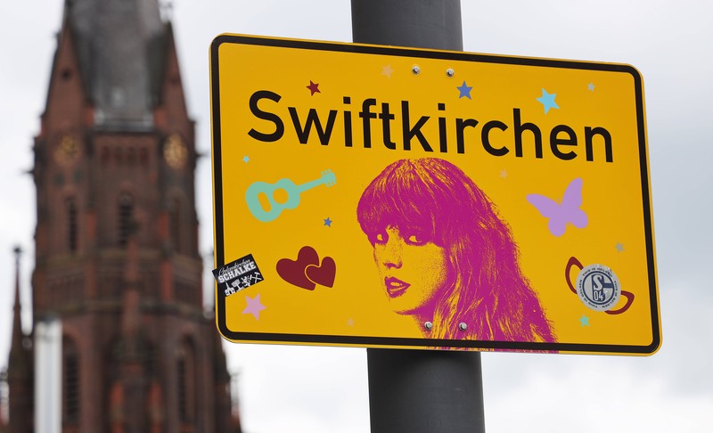 Ein Ortsschild mit der Aufschrift 'Swiftkirchen' statt 'Gelsenkirchen', daneben ein Porträt der Sängerin Taylor Swift, die im Juli drei Konzerte in der Arena auf Schalke gibt.